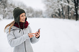 Woman in grey winter coat, red scarf, and green winter hat on snowy road with snow covered trees in the background, looking to the right and smiling with phone in hand