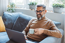 Man with grey hair, full beard, and black round glasses and wearing a brown turtleneck sitting on grey couch with laptop on his lap, holding mug in his left hand. Row of windows behind him with potted plants on windowsill